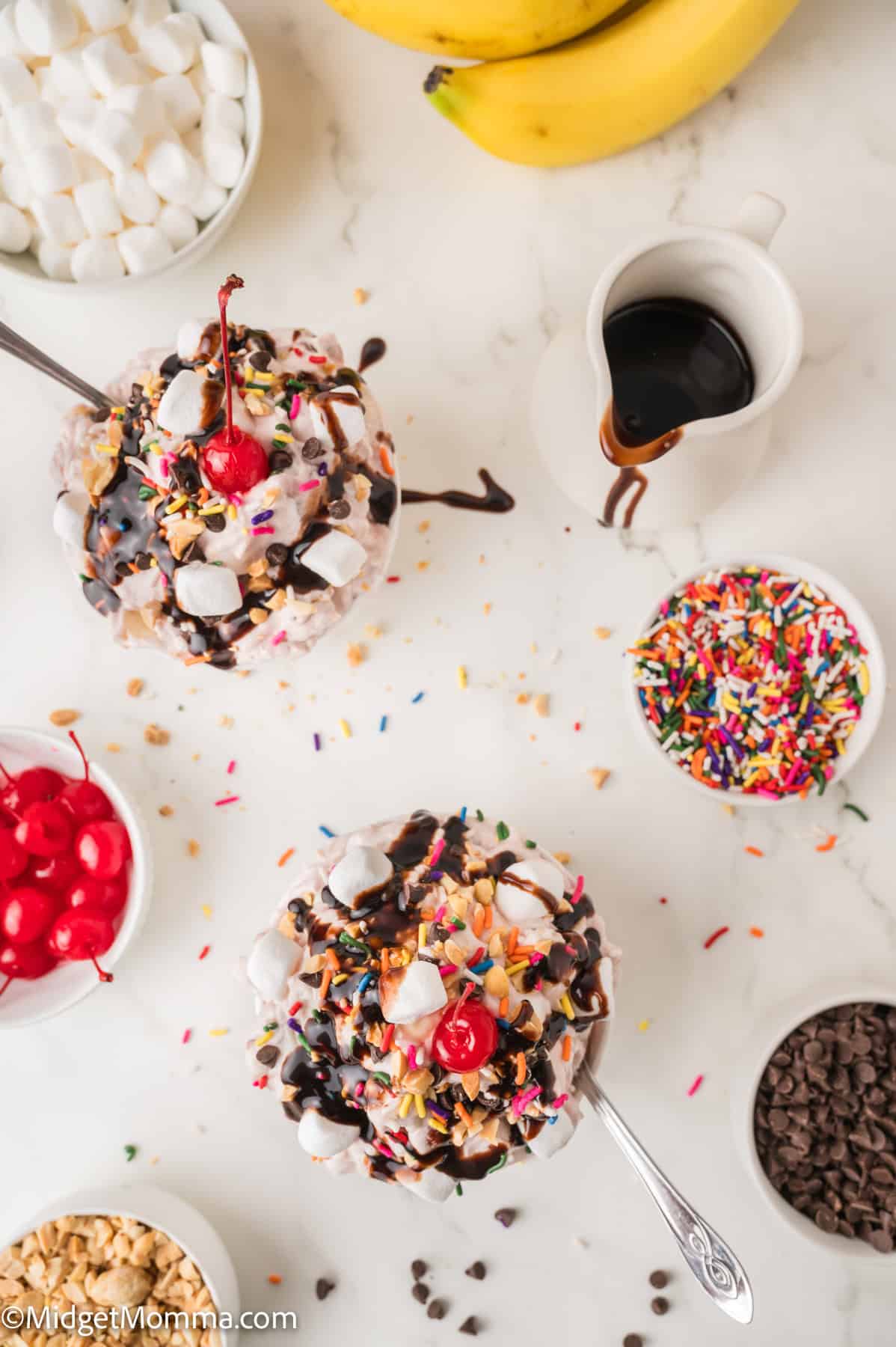 Two ice cream sundaes topped with whipped cream, sprinkles, cherries, marshmallows, and chocolate sauce, surrounded by toppings and bananas on a white surface.