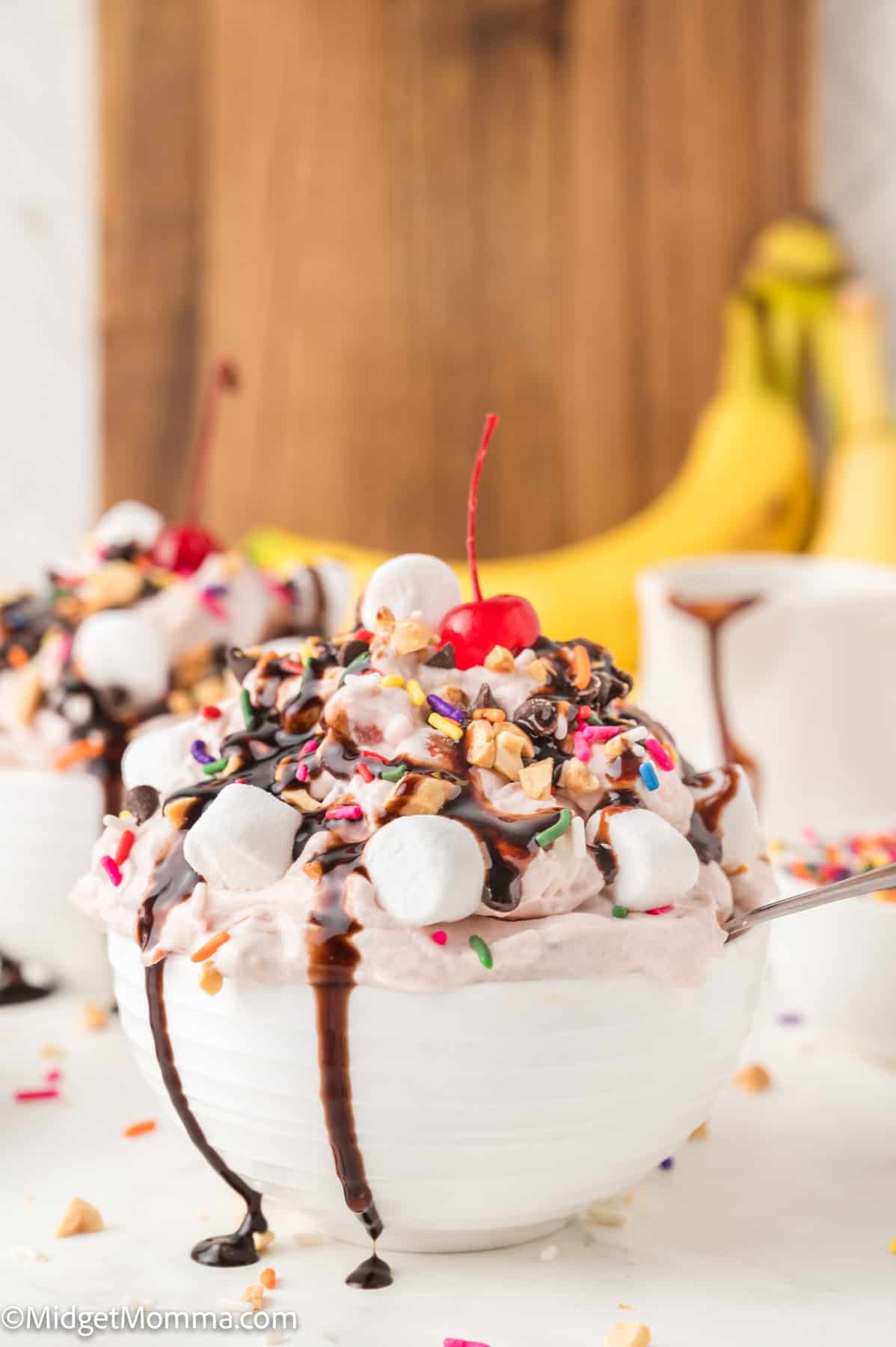 A bowl of ice cream topped with whipped cream, chocolate syrup, marshmallows, sprinkles, chopped nuts, and a cherry, with bananas and a wooden board in the background.