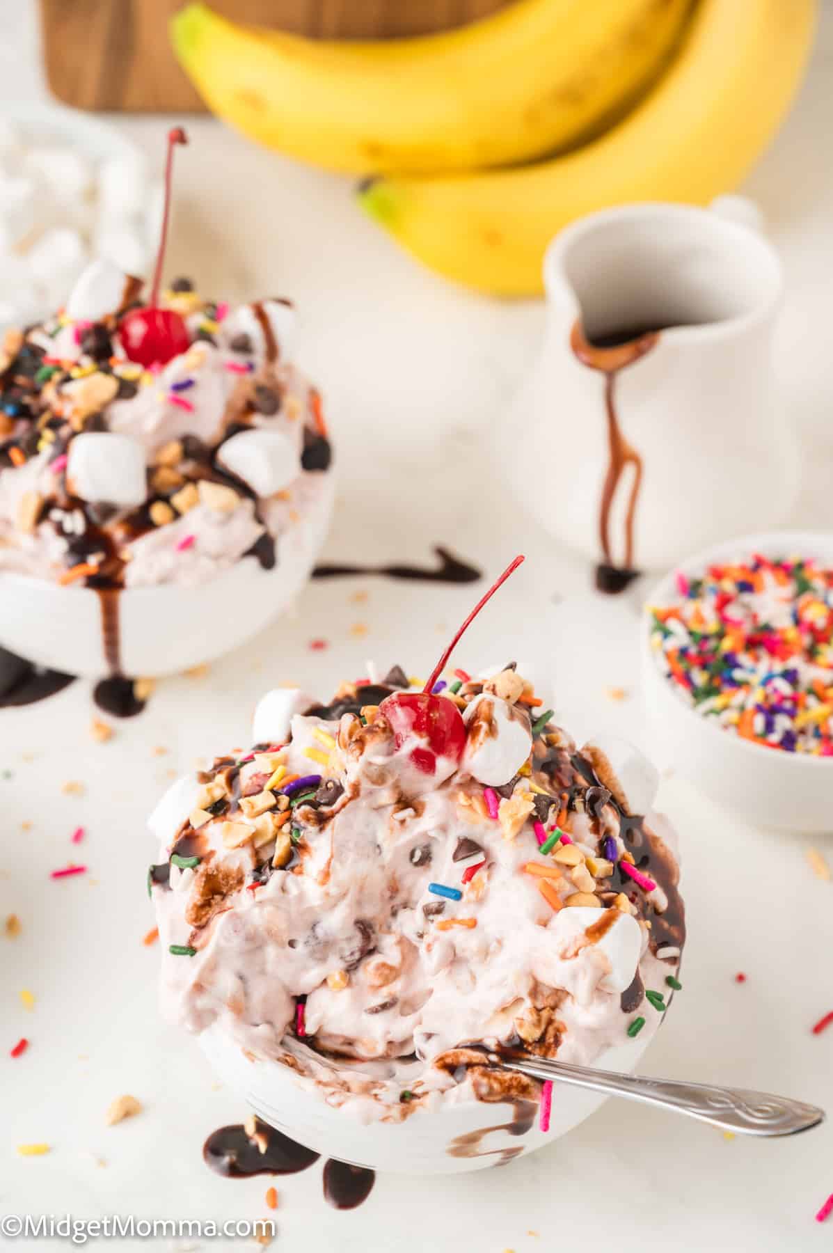 Two bowls of ice cream topped with chocolate syrup, sprinkles, nuts, marshmallows, and a cherry, with bananas and a small pitcher in the background.