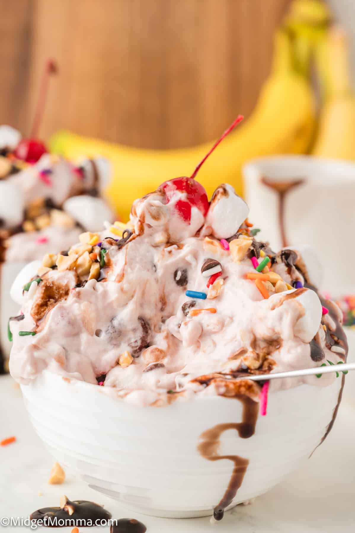 A bowl of banana split ice cream topped with whipped cream, chocolate syrup, chopped nuts, sprinkles, marshmallows, and a cherry, with bananas in the background.