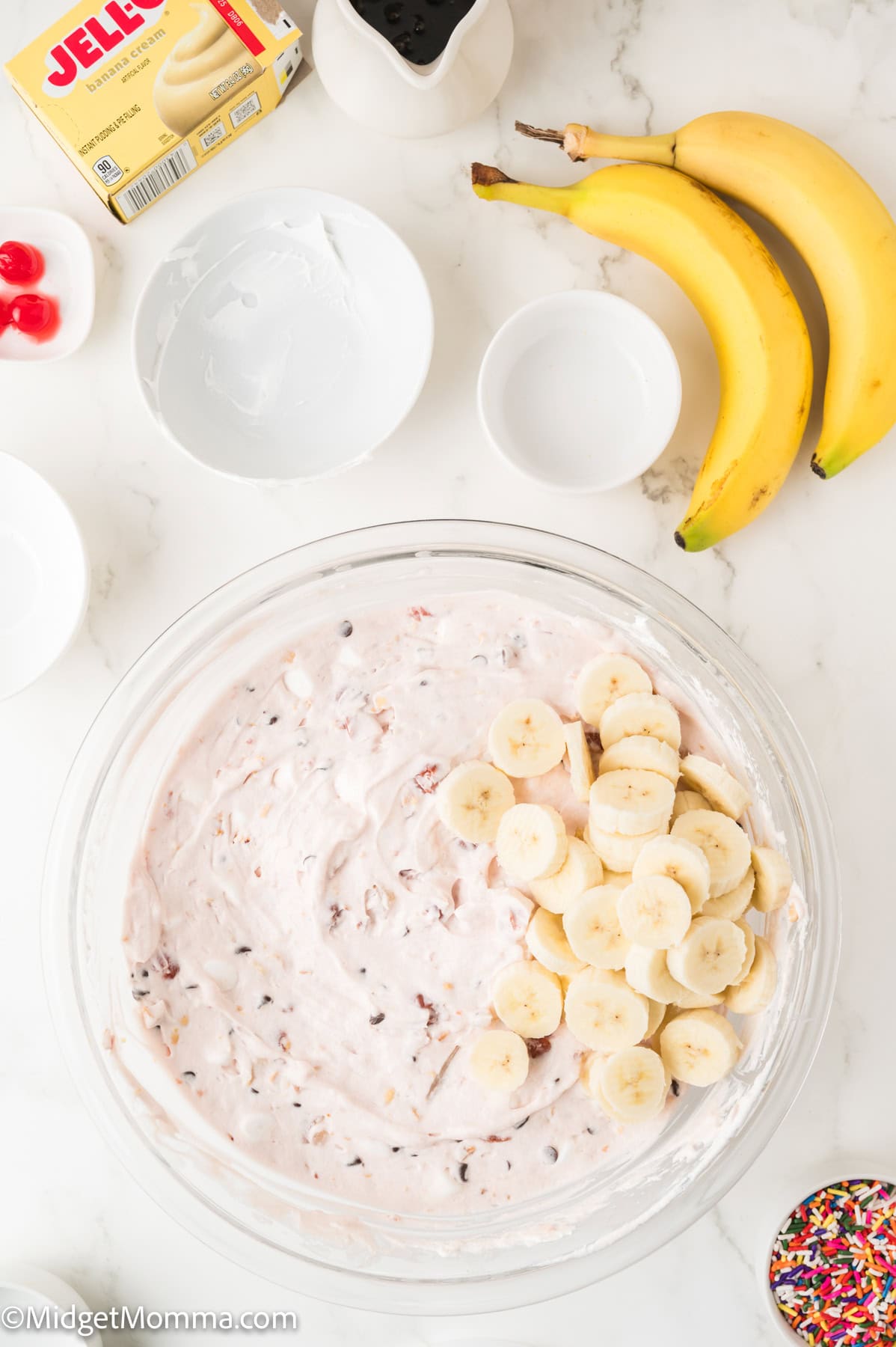 A bowl of creamy dessert mixture with banana slices, surrounded by bananas, cherries, Jell-O pudding box, and small bowls on a white surface.