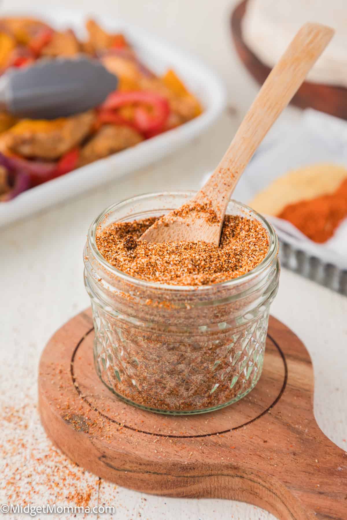 A small glass jar filled with a spice blend sits on a wooden coaster, with a wooden spoon resting inside. A tray of food is blurred in the background.
