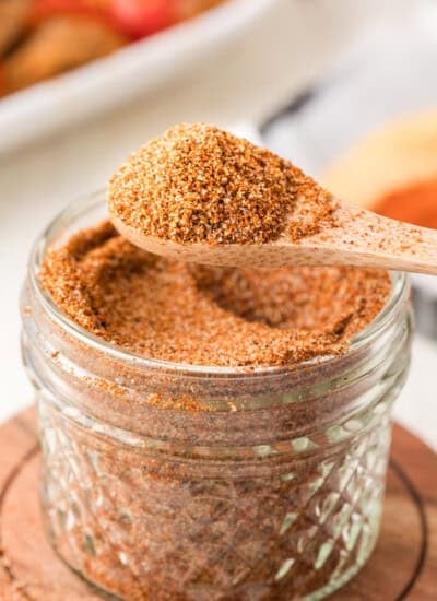 A wooden spoon holds a scoop of taco seasoning above a small glass jar filled with more taco seasoning.