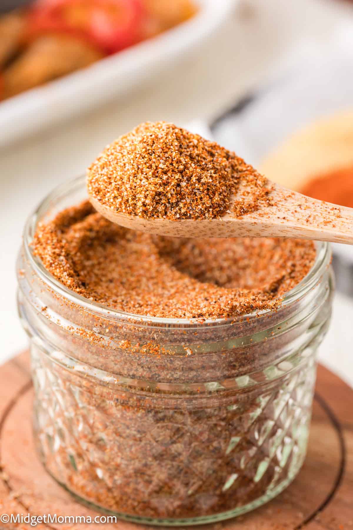 A wooden spoon holds a scoop of taco seasoning above a small glass jar filled with more taco seasoning.