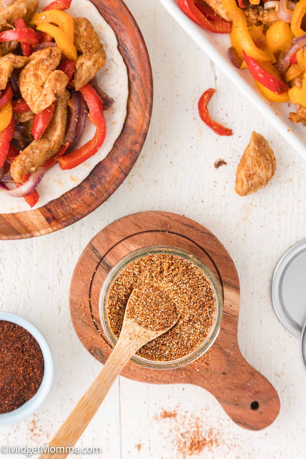 Top-down view of fajita filling with bell peppers and chicken on a tortilla, a jar of seasoning with a spoon, and additional filling in a white dish on a white surface.