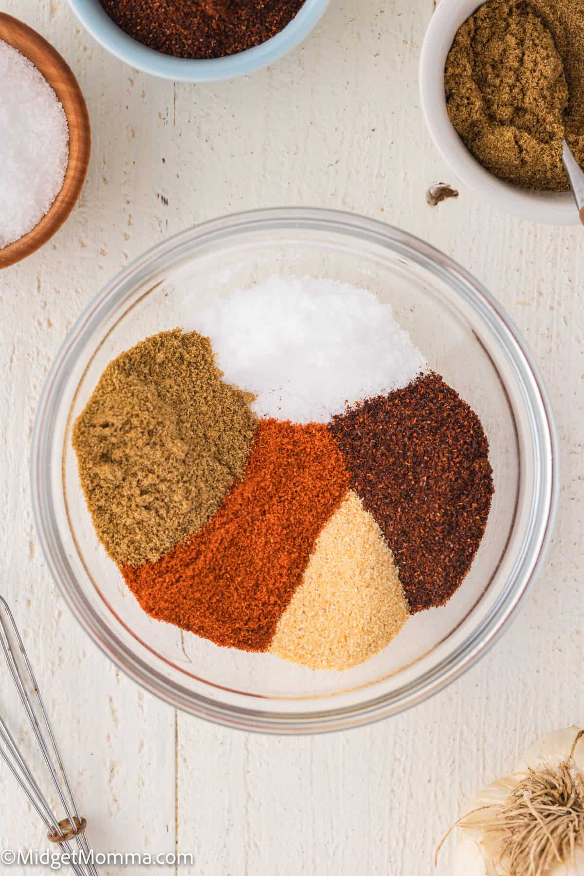 A glass bowl containing separate piles of ground spices including salt, paprika, chili powder, garlic powder, and cumin on a white surface.