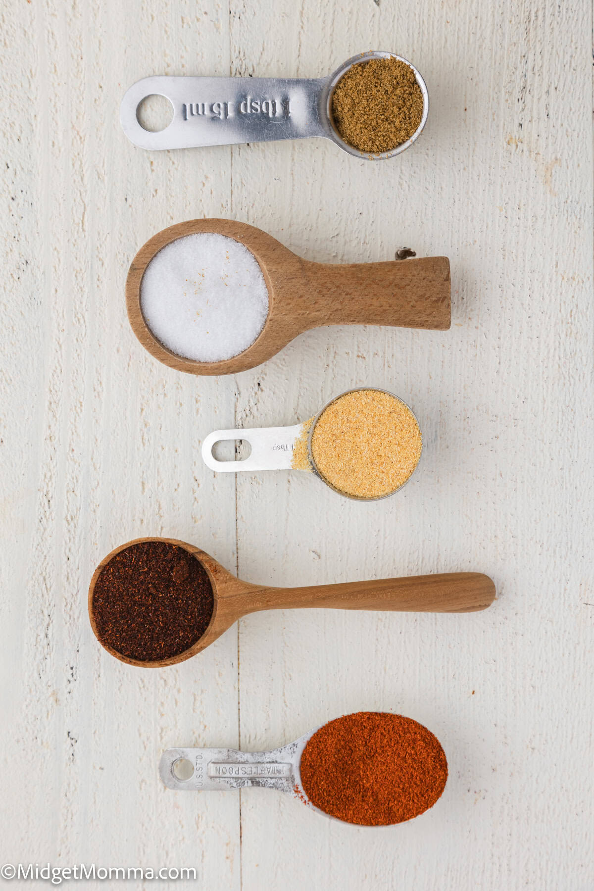 Five different dried spices and seasonings are arranged in measuring spoons and wooden scoops on a light wooden surface.