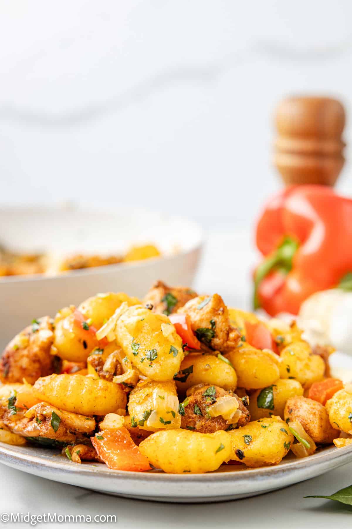 A plate of gnocchi mixed with diced chicken, red bell peppers, and herbs sits on a white table with a pepper grinder and vegetables in the background.
