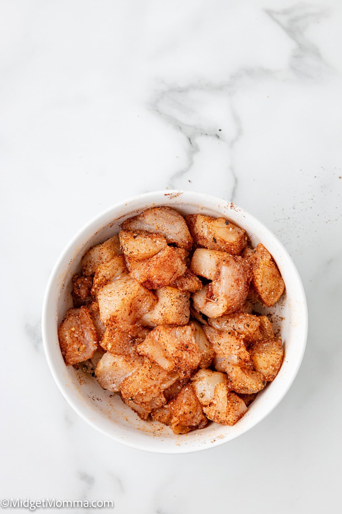 A white bowl filled with raw, diced chicken pieces coated in seasoning sits on a white marble surface.