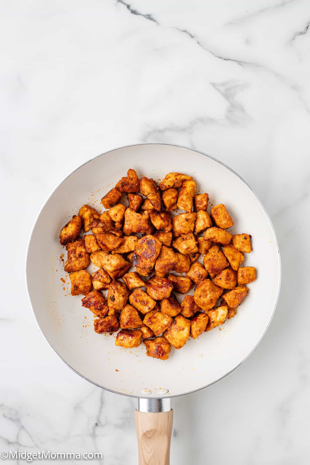 A white frying pan with cooked, seasoned chicken pieces on a marble countertop.