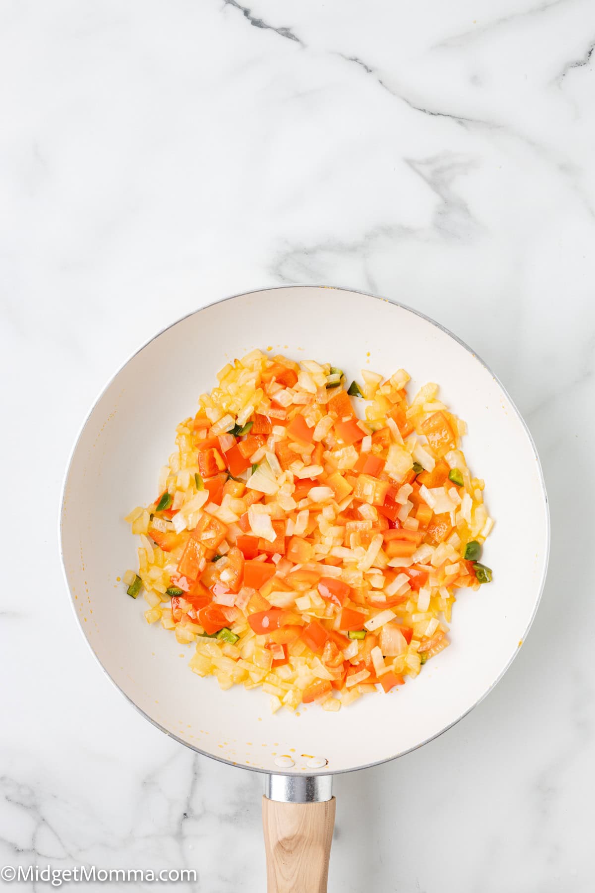 A white frying pan with diced onions, tomatoes, and green peppers cooking on a white marble surface.