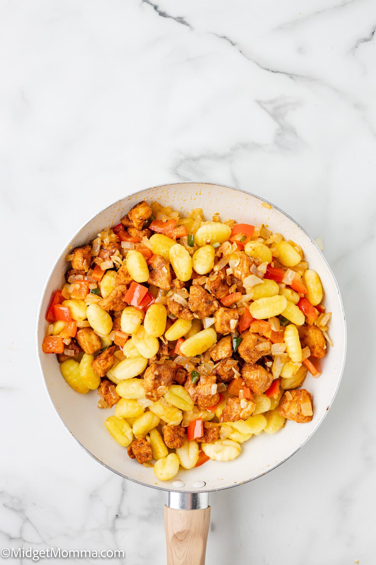 A frying pan with cooked gnocchi, diced red bell peppers, and browned pieces of sausage on a white marble surface.