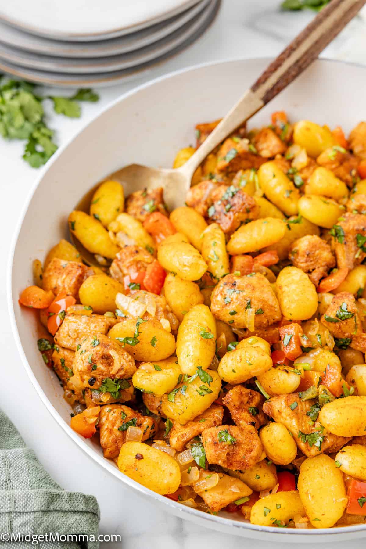 A bowl of cooked gnocchi mixed with diced chicken, carrots, and herbs, with a serving spoon resting inside.