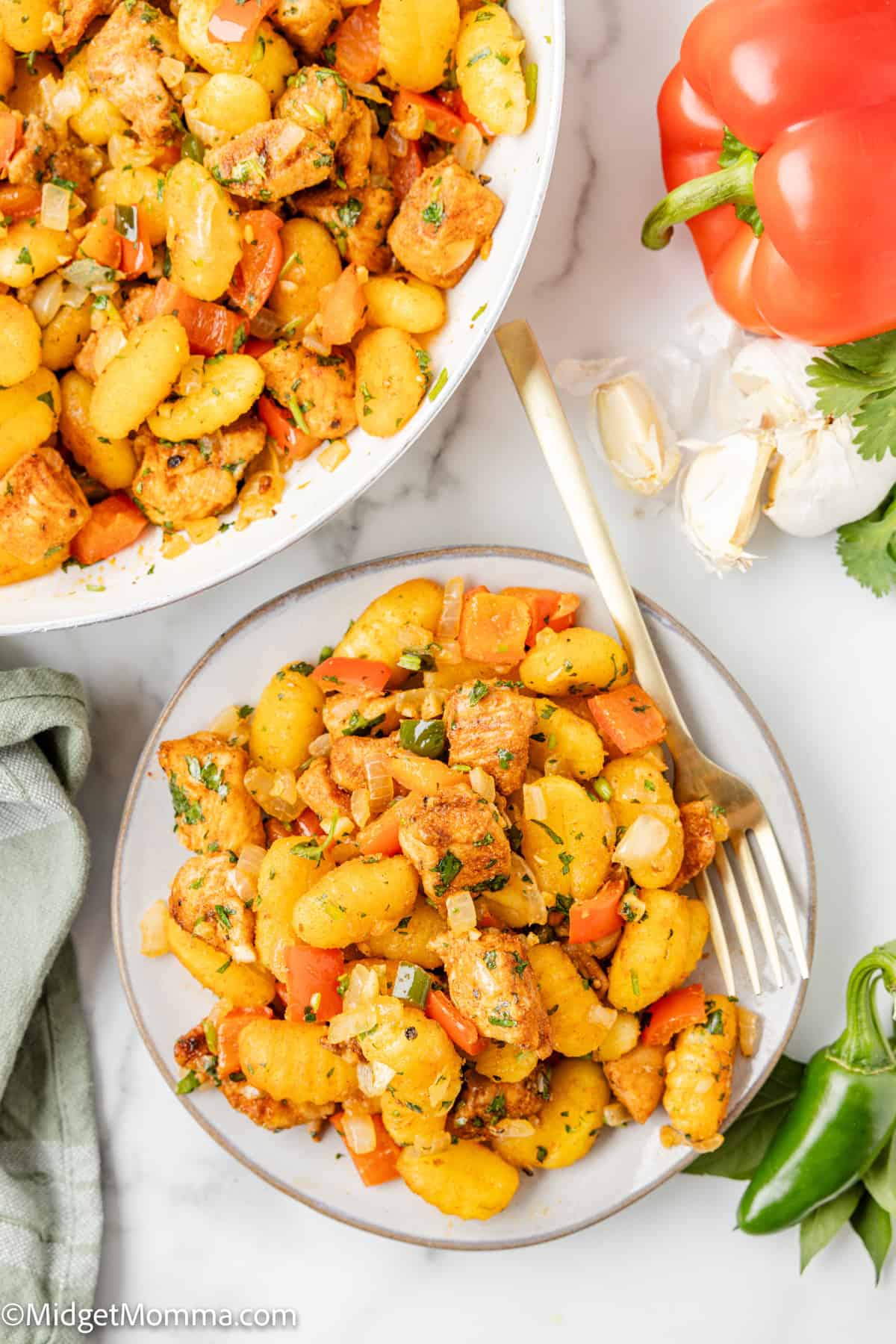 A plate of gnocchi mixed with chunks of chicken, diced red bell pepper, and herbs sits next to a skillet of the same dish, fresh vegetables, and a fork.
