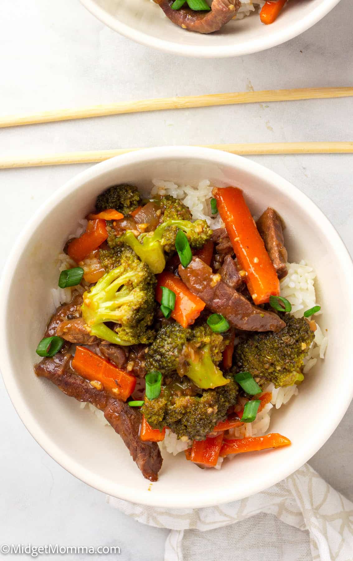 A bowl of beef stir-fry with broccoli, carrots, and green onions served over white rice, with chopsticks placed above the bowl.