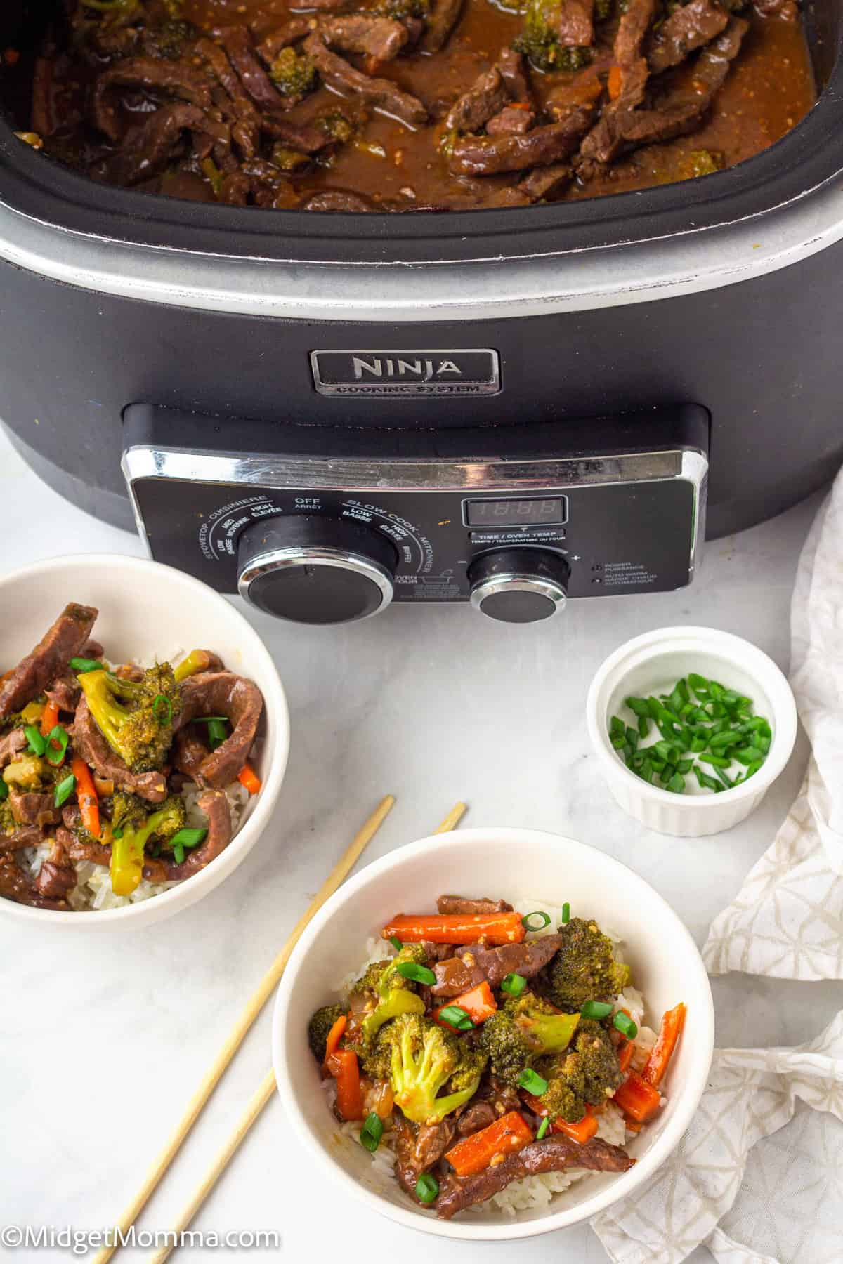 A slow cooker filled with beef and vegetables sits behind two bowls of beef stir-fry served over rice, with a small bowl of chopped green onions on the side.
