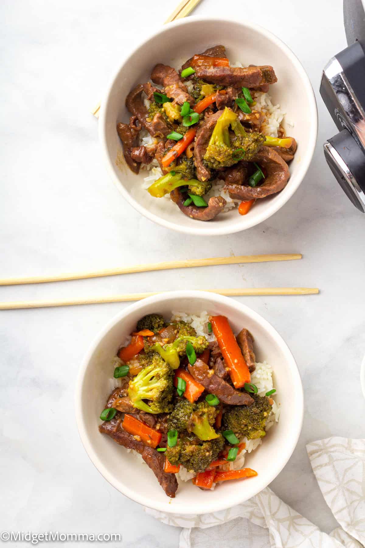 Two bowls of beef stir-fry with broccoli, carrots, and green onions served over white rice, with chopsticks beside them on a white surface.