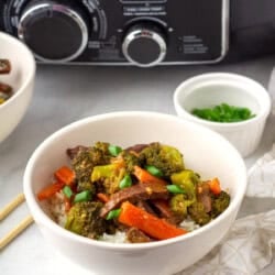 A bowl of beef stir-fry with broccoli, carrots, and rice is placed on a table with chopsticks, a small bowl of green onions, and a slow cooker in the background.