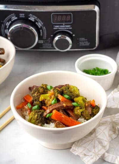 A bowl of beef stir-fry with broccoli, carrots, and rice is placed on a table with chopsticks, a small bowl of green onions, and a slow cooker in the background.