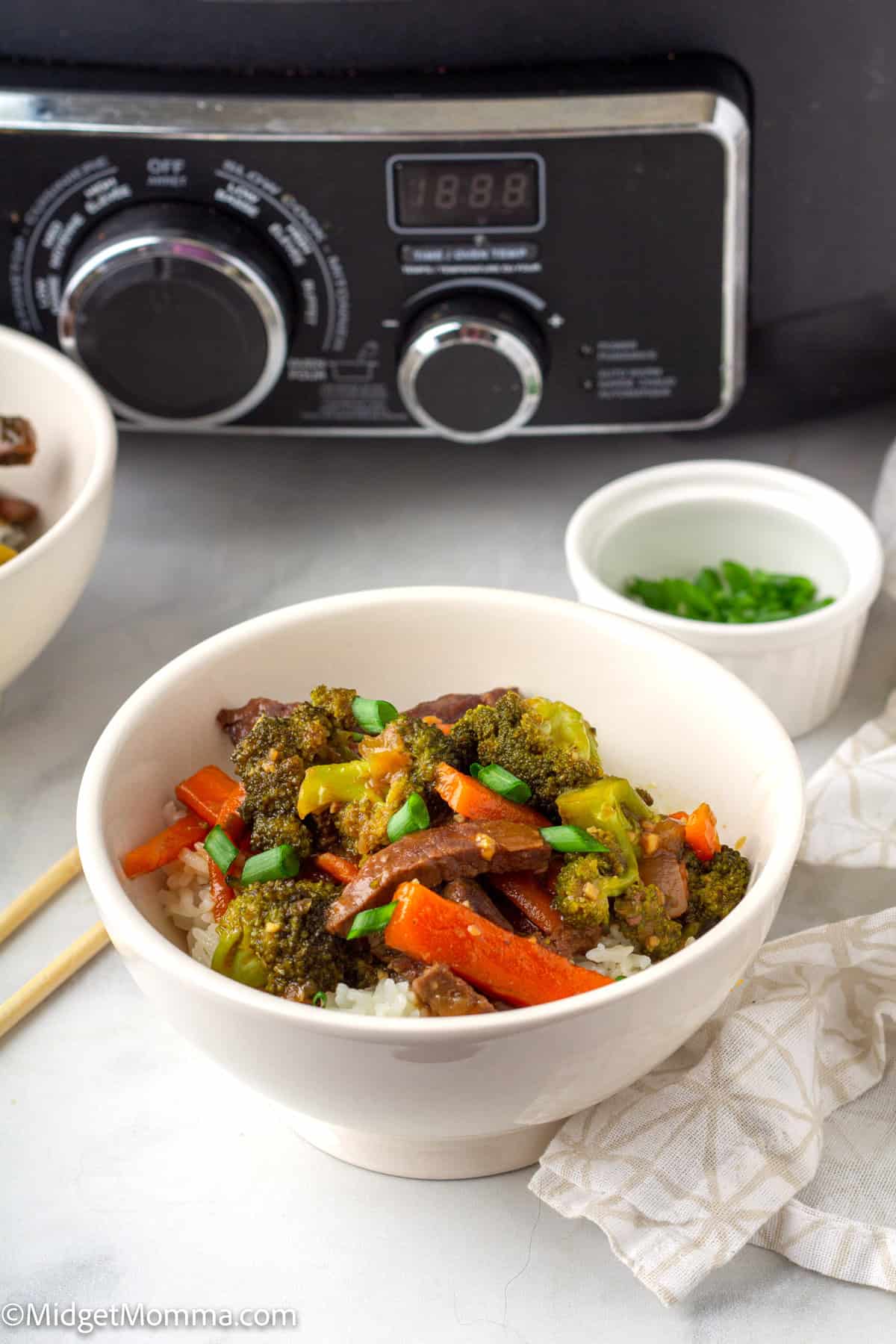 A bowl of beef stir-fry with broccoli, carrots, and rice is placed on a table with chopsticks, a small bowl of green onions, and a slow cooker in the background.