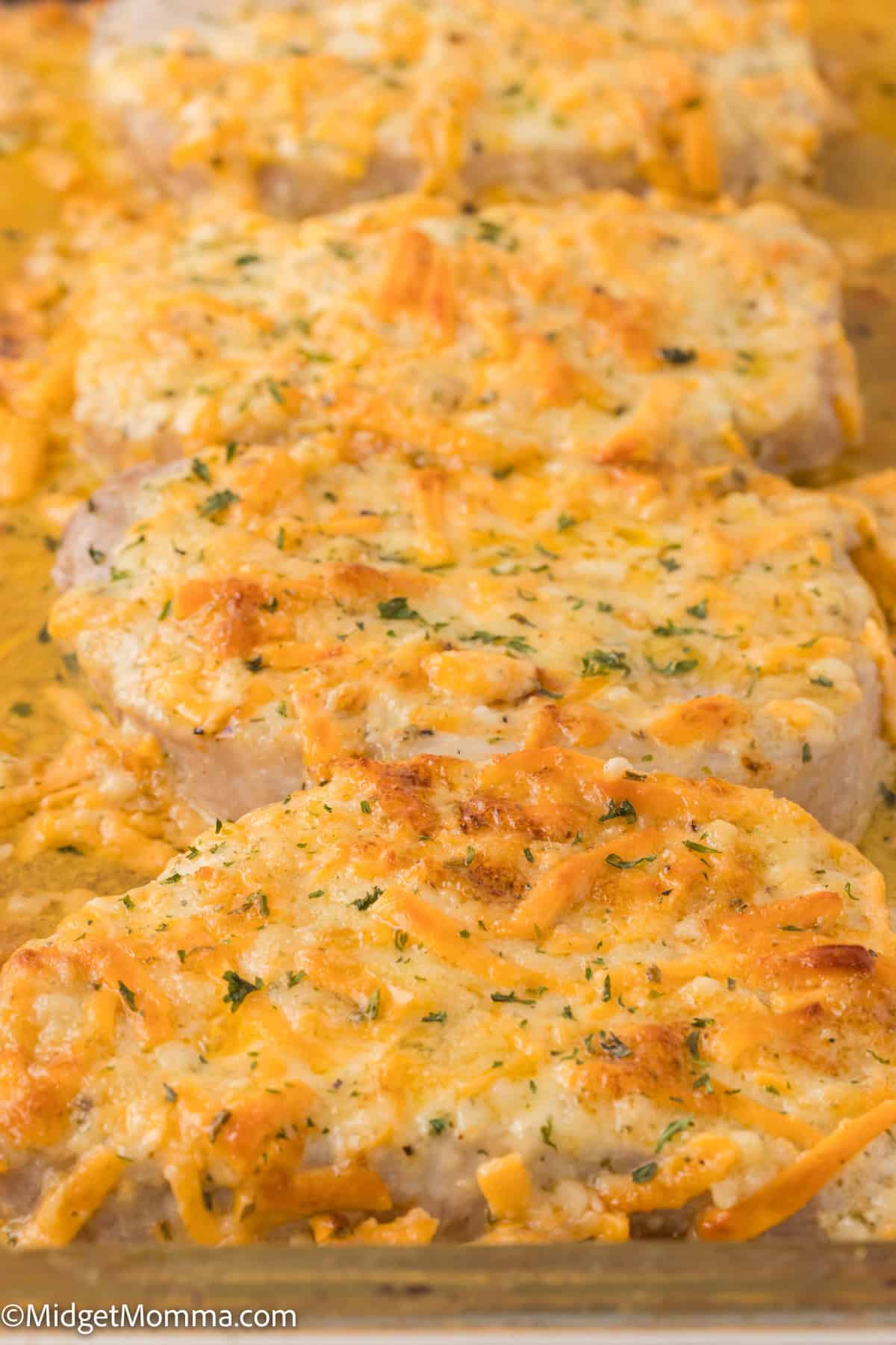 Baked pork chops topped with melted cheddar cheese and herbs, arranged in a row on a baking sheet.