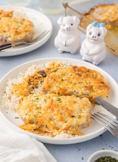 Two baked, cheese-covered chicken breasts served over white rice on a white plate, with a fork and knife, and pig-shaped salt and pepper shakers in the background.