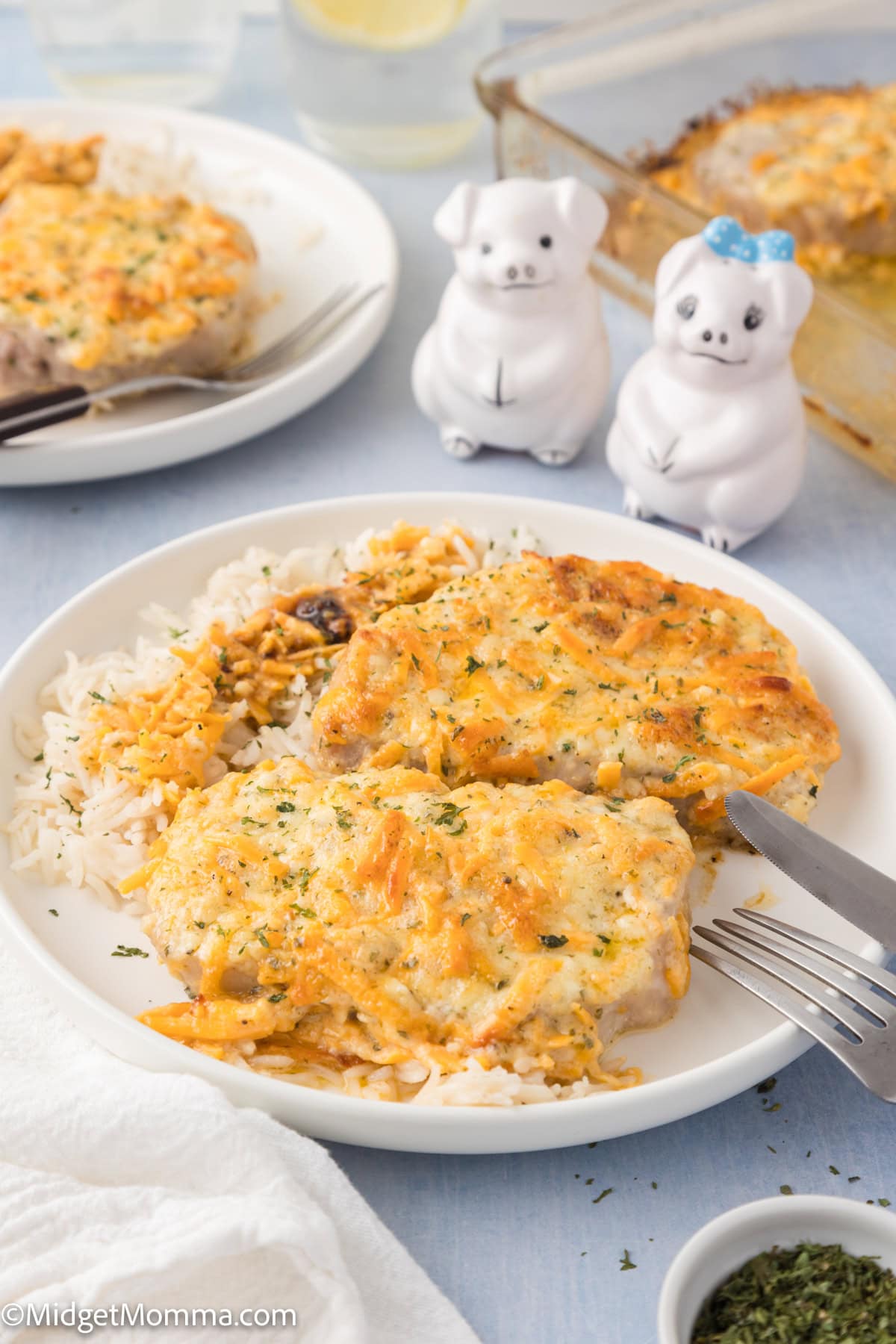 Two baked, cheese-covered chicken breasts served over white rice on a white plate, with a fork and knife, and pig-shaped salt and pepper shakers in the background.