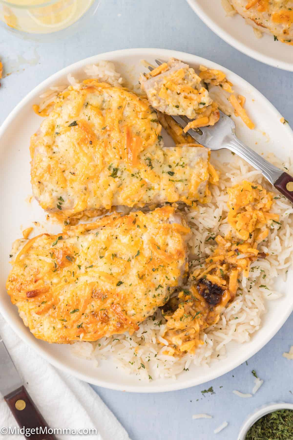 Two cheesy baked pork chops served over white rice on a white plate, with a fork holding a bite-sized piece of pork and rice.