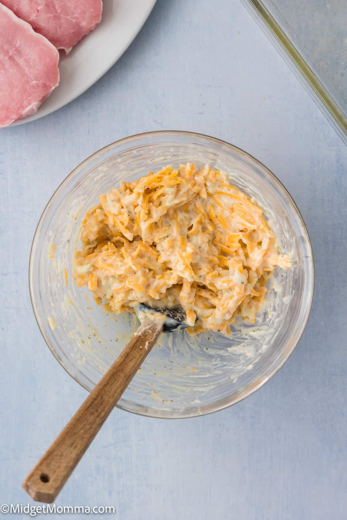 A glass bowl with a mixture of shredded cheese, grated potatoes, and creamy sauce, next to a plate with raw pork chops.