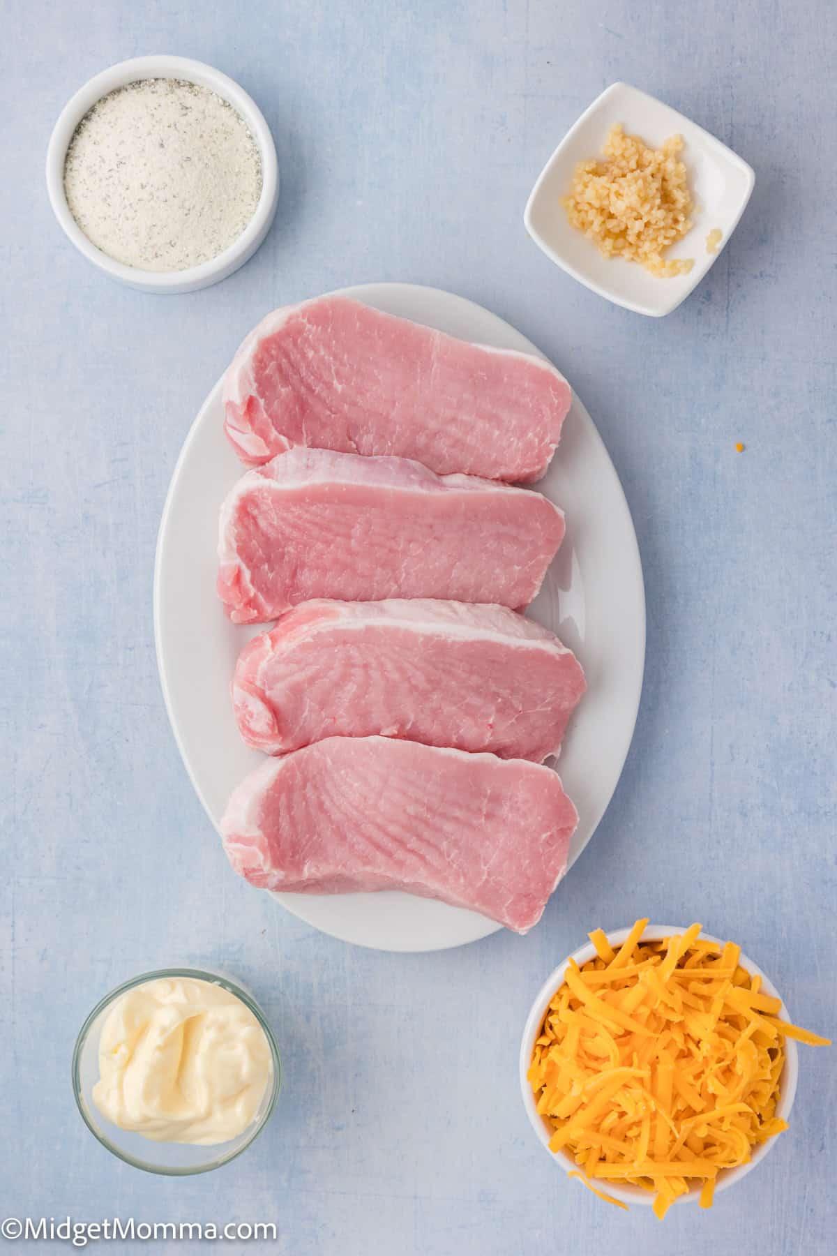 Four raw pork chops are arranged on a white plate, surrounded by small bowls containing shredded cheese, mayonnaise, ranch seasoning, and minced garlic on a light blue surface.