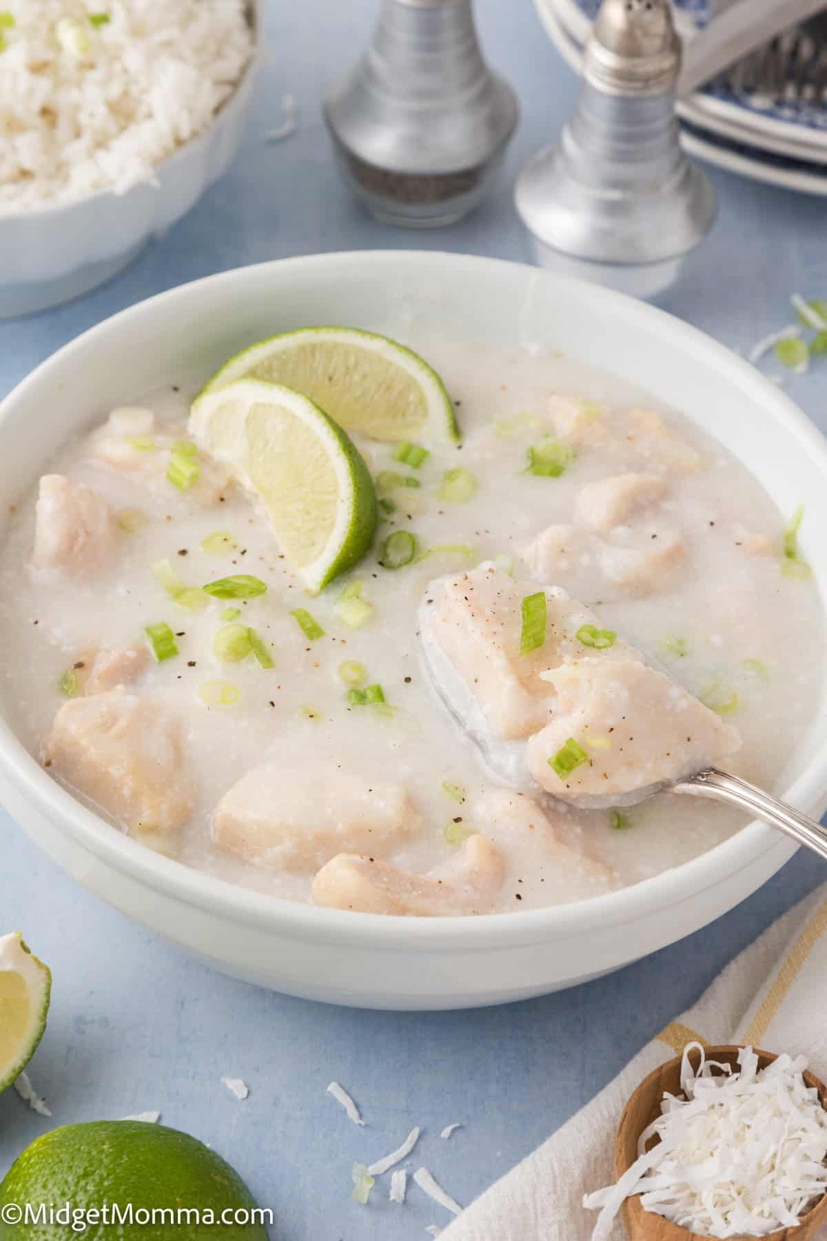 A bowl of creamy chicken soup garnished with lime wedges and chopped green onions, with a spoon inside. Rice and seasonings are visible in the background.