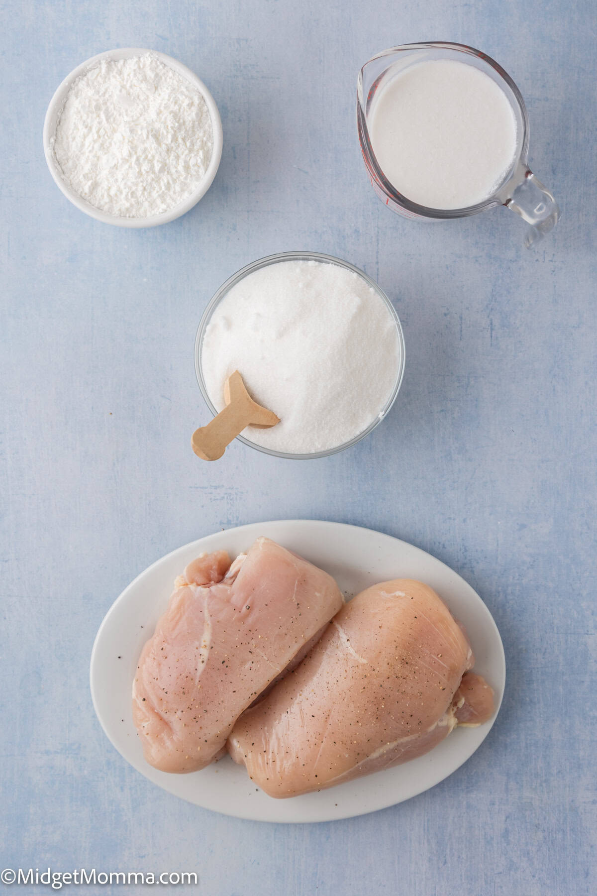 Raw chicken breasts on a plate, a bowl of flour, a bowl of white powder with a scoop, and a measuring cup of milk arranged on a light blue surface.