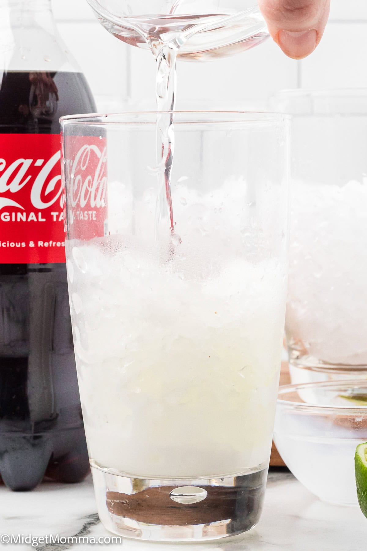 A glass filled with ice and a clear liquid being poured in, with a bottle of Coca-Cola and bowls of ice and limes in the background.