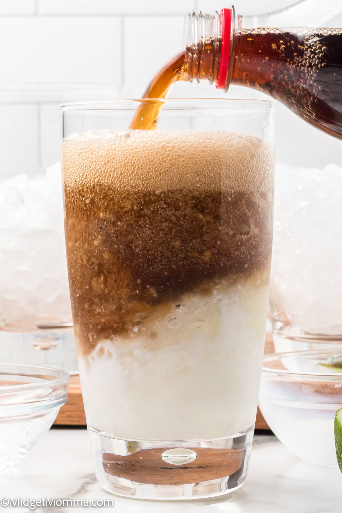 A glass filled with crushed ice and a creamy white liquid is being topped with a dark soda poured from a plastic bottle.