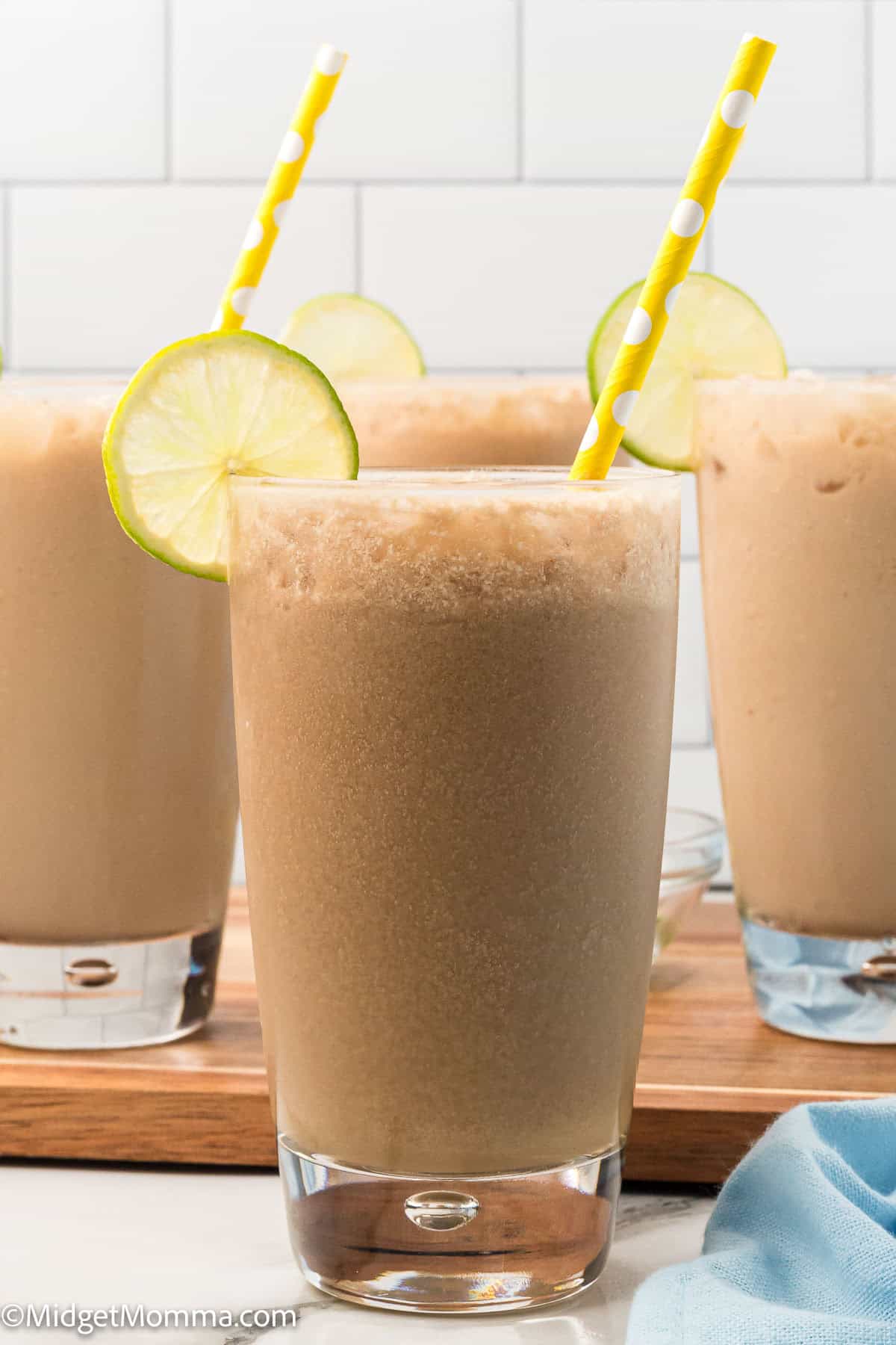 Four glasses of a frothy brown beverage with ice, garnished with lime slices and yellow striped straws, placed on a wooden tray.