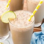 Four glasses of iced coffee with frothy tops, yellow polka dot straws, and lime slices on the rims, set on a table with a blue cloth and extra lime slices.