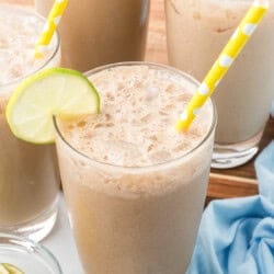 Four glasses of iced coffee with frothy tops, yellow polka dot straws, and lime slices on the rims, set on a table with a blue cloth and extra lime slices.