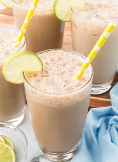 Four glasses of iced coffee with frothy tops, yellow polka dot straws, and lime slices on the rims, set on a table with a blue cloth and extra lime slices.