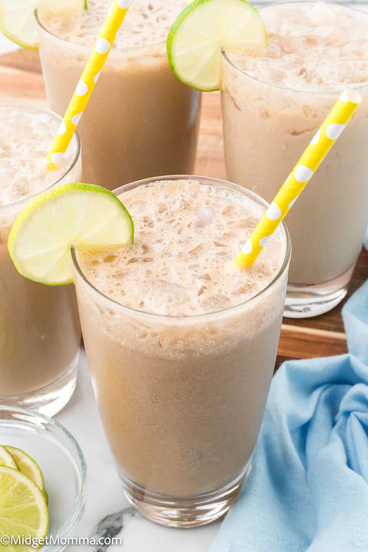 Four glasses of iced coffee with frothy tops, yellow polka dot straws, and lime slices on the rims, set on a table with a blue cloth and extra lime slices.