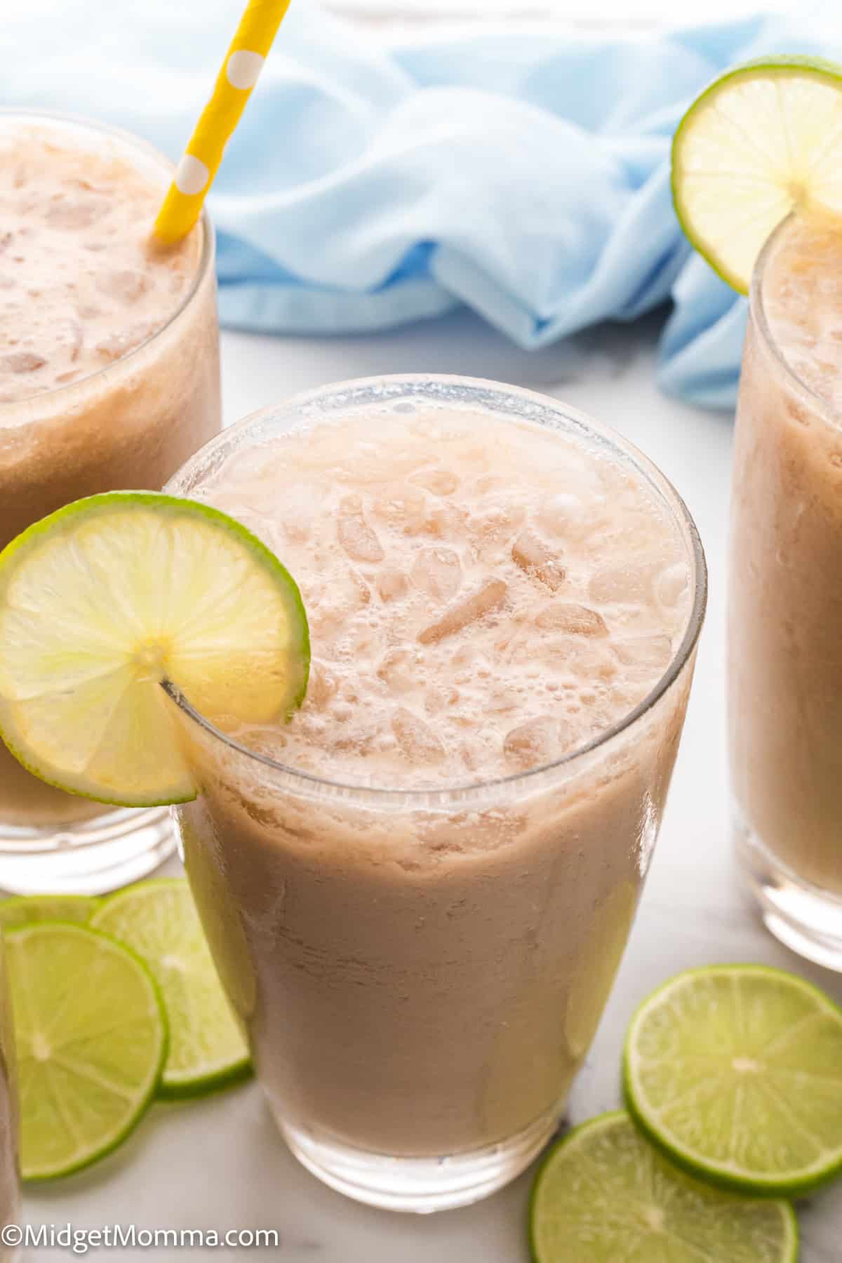 Three glasses of iced drink garnished with lime slices, one with a yellow polka dot straw, surrounded by lime slices on a white surface.