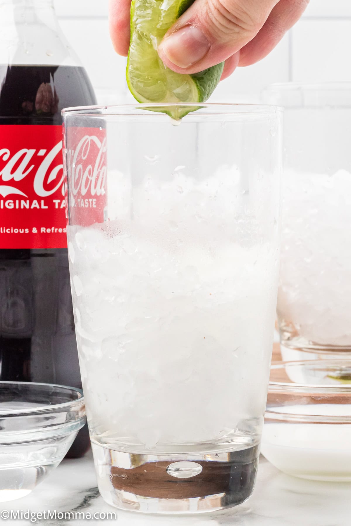 A hand squeezes a lime over a glass filled with crushed ice, with a bottle of Coca-Cola and a bowl of more ice nearby.