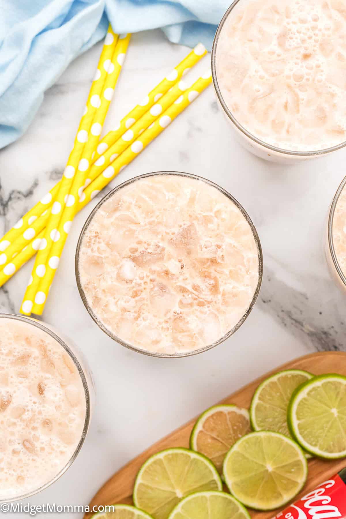 Four glasses of iced creamy beverage on a marble surface, with yellow polka dot straws, a blue cloth, and sliced limes on a wooden board.