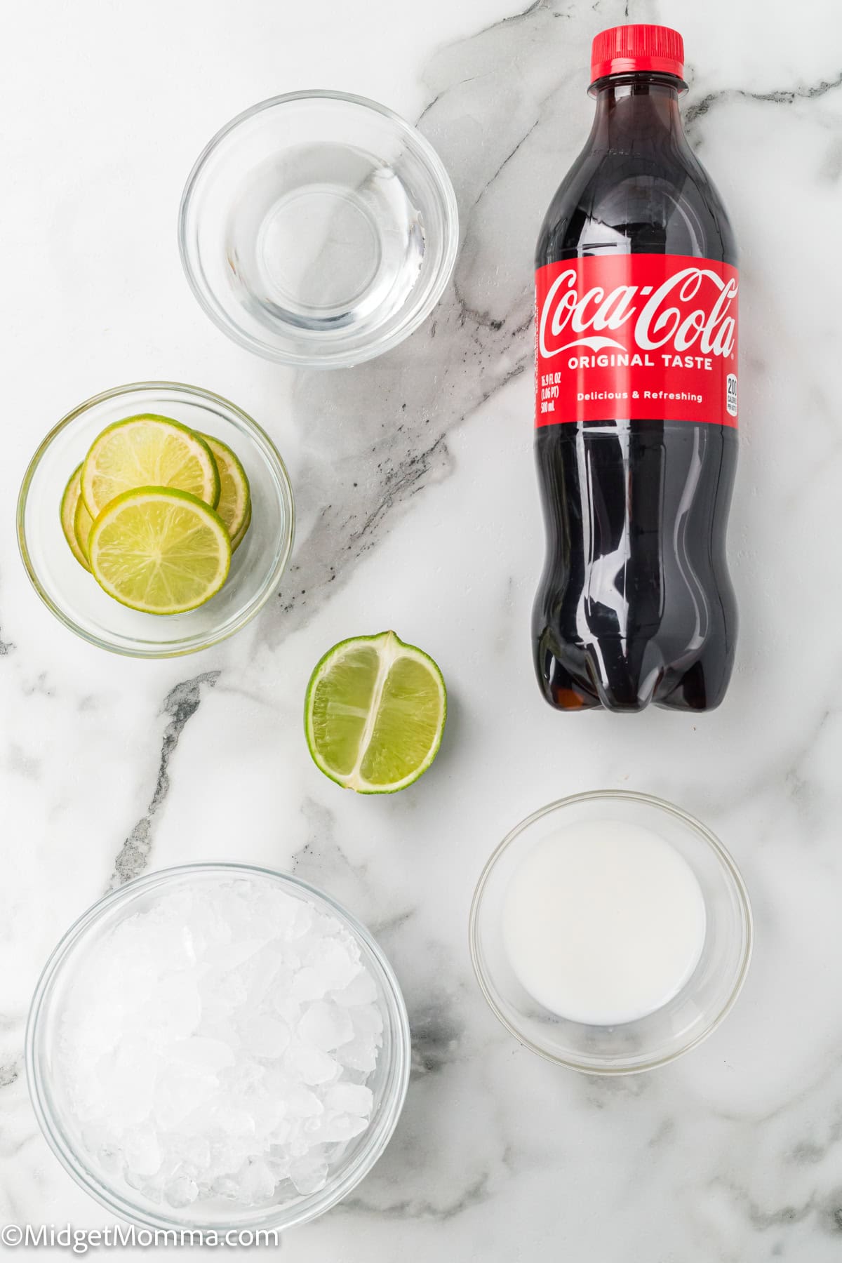 A bottle of Coca-Cola, a bowl of ice, a bowl of lime slices, a halved lime, a bowl of sugar, and a glass of water on a marble surface.