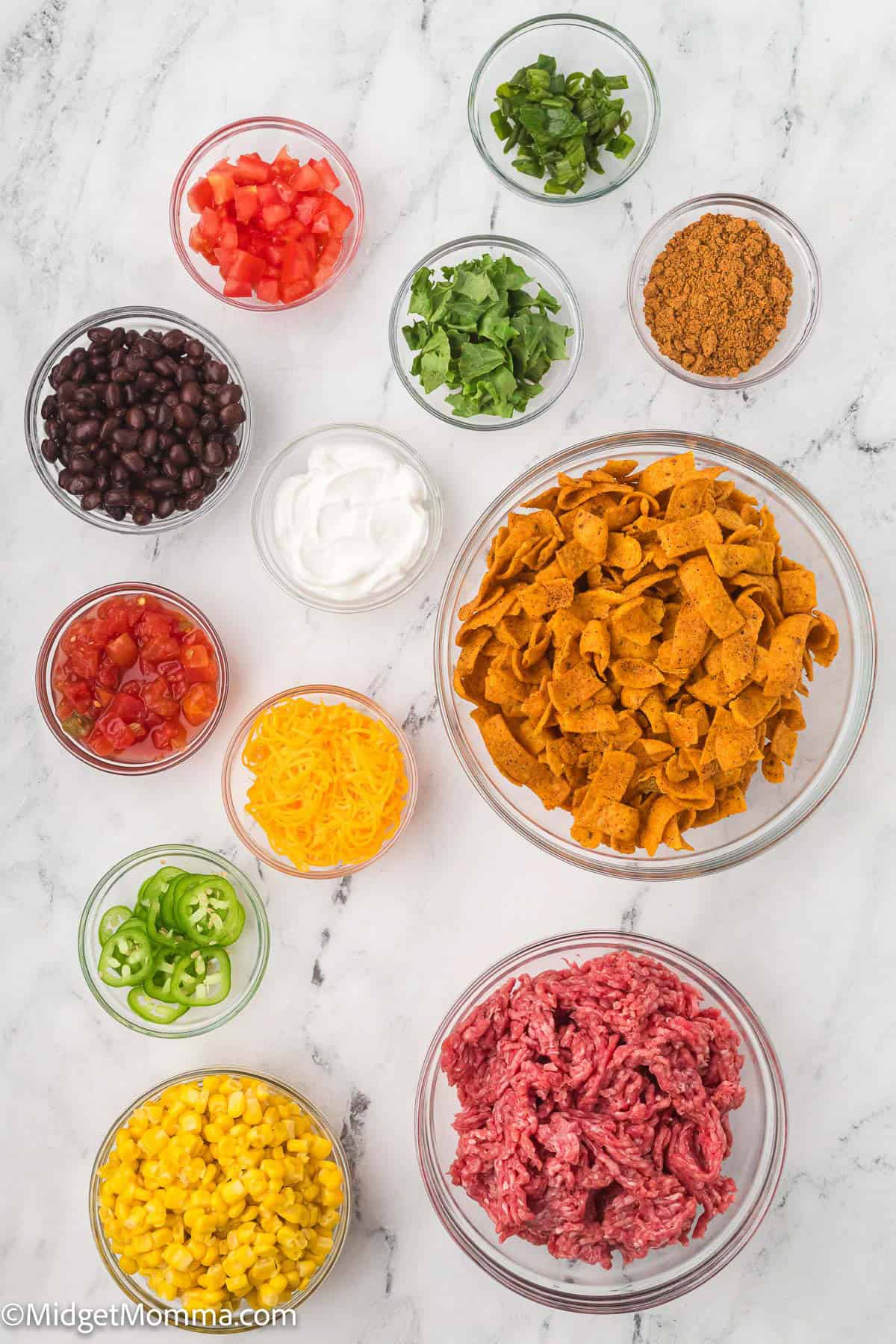 Bowls of chopped tomatoes, cilantro, green onions, taco seasoning, black beans, sour cream, Fritos, shredded cheddar, canned tomatoes, jalapeños, corn, and raw ground beef on a marble surface.