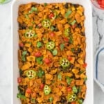 A rectangular white baking dish filled with a casserole topped with corn chips, black beans, sliced jalapeños, chopped tomatoes, and fresh herbs.