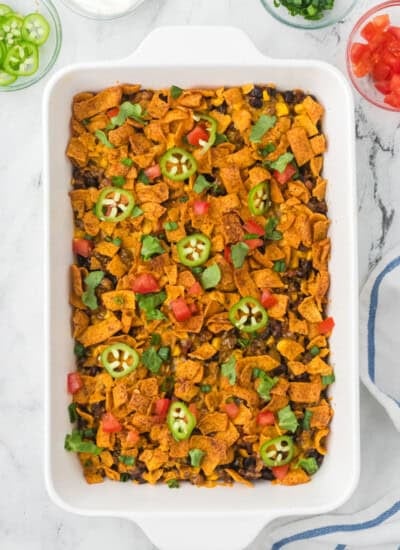 A rectangular white baking dish filled with a casserole topped with corn chips, black beans, sliced jalapeños, chopped tomatoes, and fresh herbs.