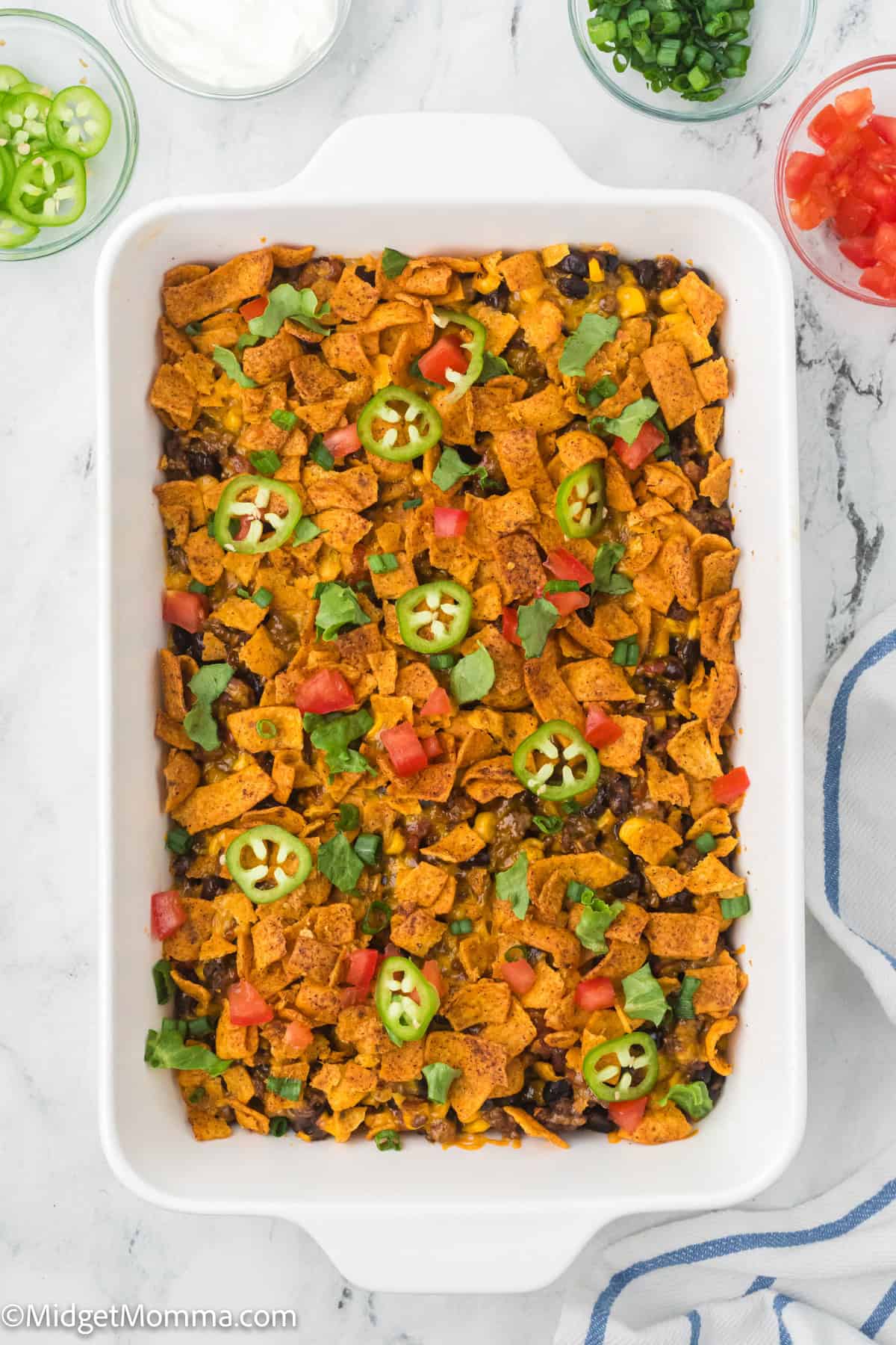 A rectangular white baking dish filled with a casserole topped with corn chips, black beans, sliced jalapeños, chopped tomatoes, and fresh herbs.