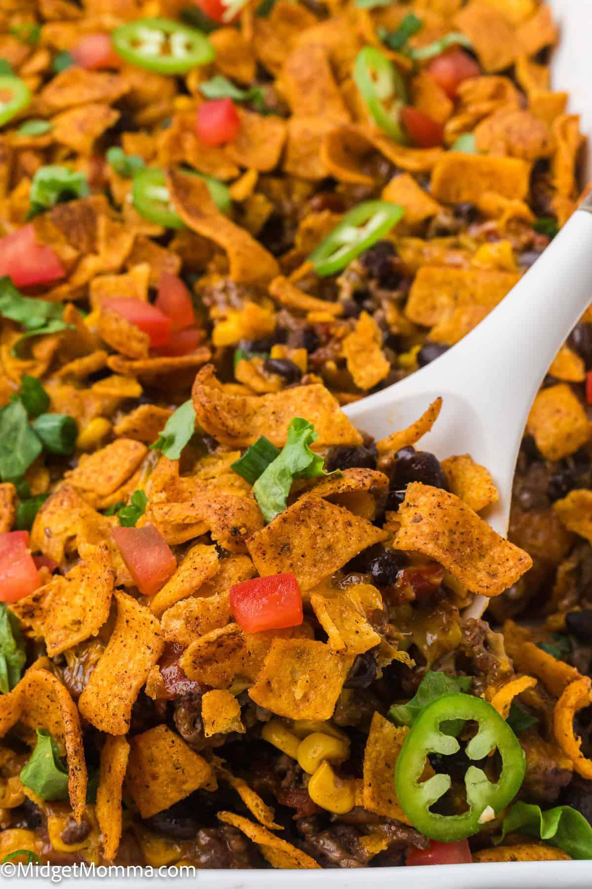 A close-up of a casserole topped with corn chips, sliced jalapeños, diced tomatoes, and chopped lettuce, being served with a white spoon.