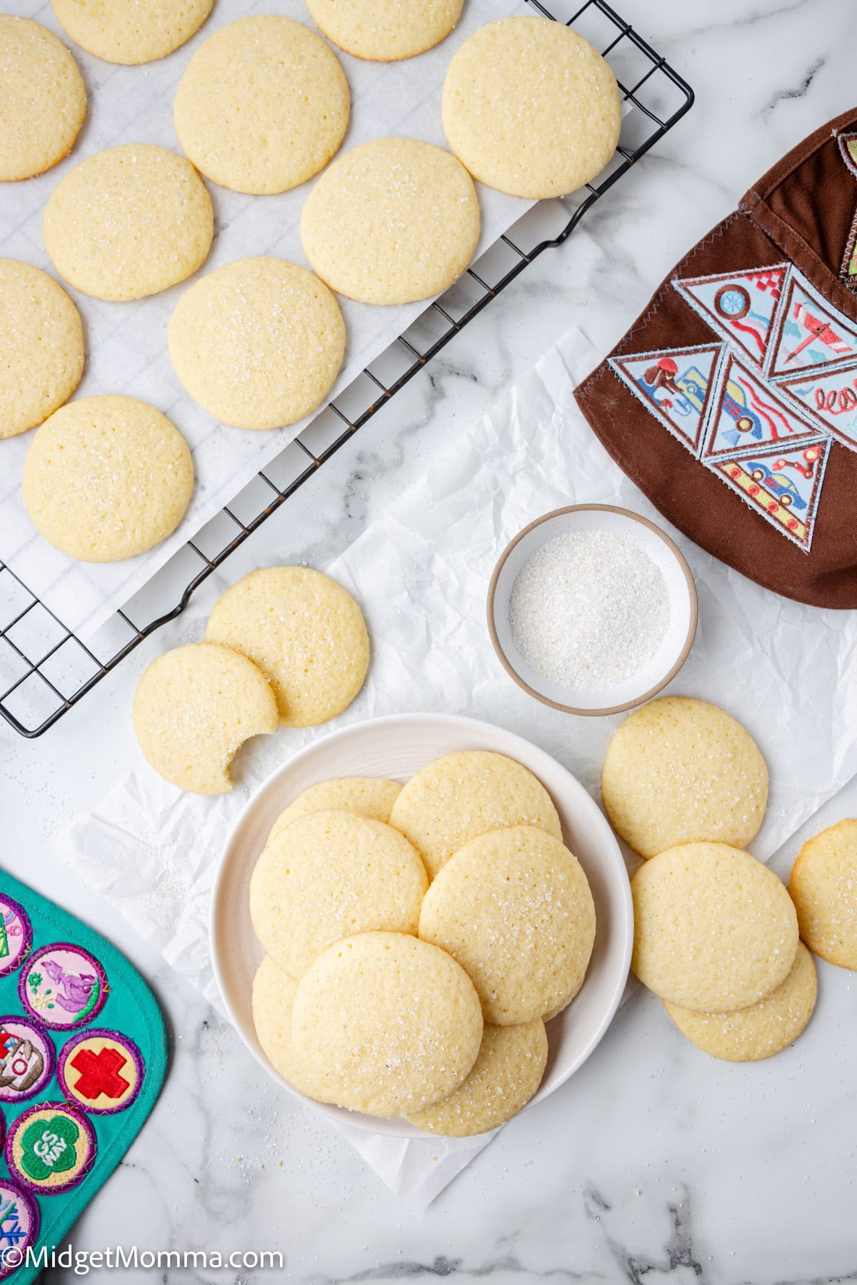 Round sugar cookies are arranged on a cooling rack and a plate, with a small bowl of sugar nearby and a decorated pouch and a patch-covered item visible in the scene.