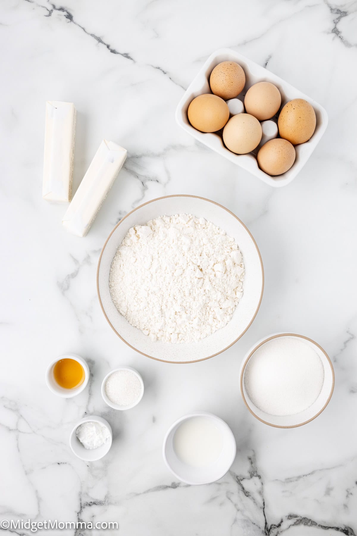 Baking ingredients on a marble surface: six eggs, two sticks of butter, a bowl of flour, sugar, milk, vanilla, baking powder, and salt in small bowls.